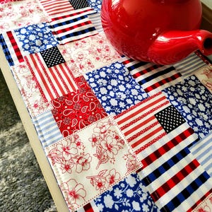 May include: A patchwork table runner with a patriotic theme. The runner features squares with American flags, red and white stripes, floral patterns, and paisley designs. A red teapot is in the upper right corner.