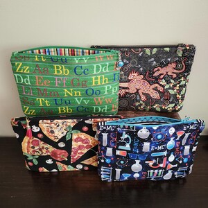 May include: Four fabric zipper pouches with various patterns. One pouch features the alphabet, another has a pizza design, a third displays a dragon, and the last one has a science theme with the formula E=MC². The pouches are on a dark wooden surface.