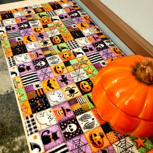 May include: A rectangular quilted table runner featuring a patchwork pattern of Halloween-themed squares. The fabric displays orange pumpkins, white ghosts, black cats, skulls, spider webs, stars, and black and white stripes in a repeating grid.
