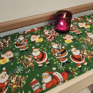 May include: A festive Christmas table runner with a green background featuring Santa Claus illustrations, snowflakes, and reindeer. A lit, dark pink candle sits on the runner. The runner is made of fabric.