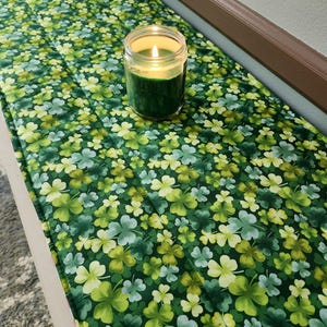 May include: A green table runner with a shamrock pattern. A lit candle in a glass jar sits on the runner.