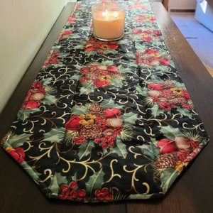 May include: A quilted table runner with a black background, featuring a repeating pattern of red berries, green holly leaves, and pinecones. A lit candle in a glass jar sits on the runner. The runner is approximately 12 inches wide.