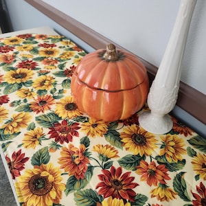 May include: A bench with a floral sunflower pattern, featuring yellow, orange, and red blooms on a cream background. An orange pumpkin-shaped ceramic container and a white vase are on the bench, creating a fall-themed display.