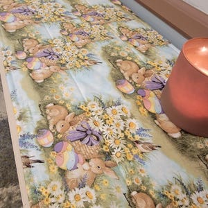 May include: Easter-themed table runner featuring a repeating pattern of bunnies, Easter eggs, and floral arrangements. The design includes pastel colors on a light blue background. A lit candle in a copper-colored holder is on the right side.