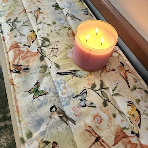 May include: A beige table runner with a floral pattern featuring butterflies, dragonflies, and birds. The runner is decorated with a vintage-inspired design and has a quilted texture. A pink candle with three wicks is sitting on the runner.