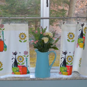 May include: Three white curtains with a black cat and flower design. The curtains are hung on a silver curtain rod. A blue pitcher with a bouquet of white flowers sits on the windowsill.