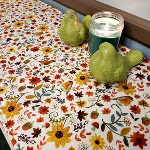 May include: A white table runner with a colorful autumn floral pattern featuring sunflowers, leaves, and birds. Two green ceramic bird figurines and a teal candle in a glass jar are on the runner.