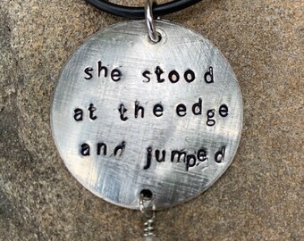 以下が含まれることがあります： 「she stood at the edge and jumped」と書かれた丸いペンダントが付いたシルバーネックレス。ペンダントはピンクの石のビーズに繋がっています。
