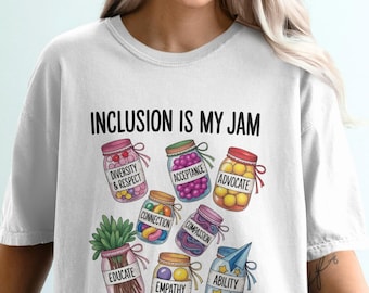 Può includere: T-shirt bianca con la scritta "INCLUSION IS MY JAM" sopra barattoli colorati con parole come "Diversità e Rispetto", "Accettazione" e "Sostenitore". La maglietta ha una vestibilità casual e rilassata.