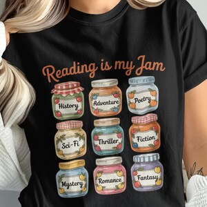 Pode incluir: Camiseta preta com a frase "Reading is my Jam" e ilustrações de potes de geleia rotulados com gêneros de livros como História, Aventura e Ficção Científica. O design é colorido e divertido.