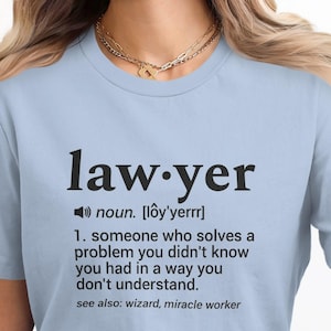 Könnte beinhalten: Hellblaues T-Shirt mit dem Wort "lawyer" in schwarzer Schrift und der Definition darunter. Das T-Shirt besteht aus einem weichen Material. Die Definition enthält die Wörter "wizard, miracle worker".