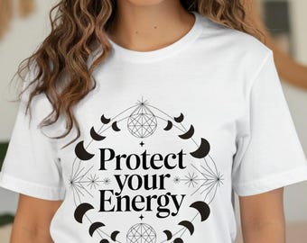 Puede incluir: Camiseta blanca con un diseño gráfico negro. El diseño presenta una forma geométrica con lunas crecientes, estrellas y las palabras "Protect your Energy". La camiseta es de cuello redondo y manga corta.