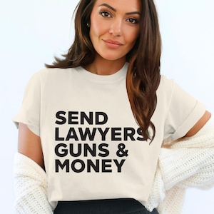 Könnte beinhalten: Cremefarbenes T-Shirt mit dem Text "SEND LAWYERS GUNS & MONEY" in fetten schwarzen Buchstaben. Das Shirt ist ein kurzärmeliger Rundhalsausschnitt. Die Person trägt eine weiße Strickjacke und eine schwarze Hose.