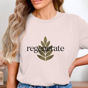 Puede incluir: Camiseta rosa claro con la palabra "regenerate" en escritura negra y un dise&ntilde;o de hojas verdes. La camiseta est&aacute; hecha de un material suave y es adecuada para ropa casual.