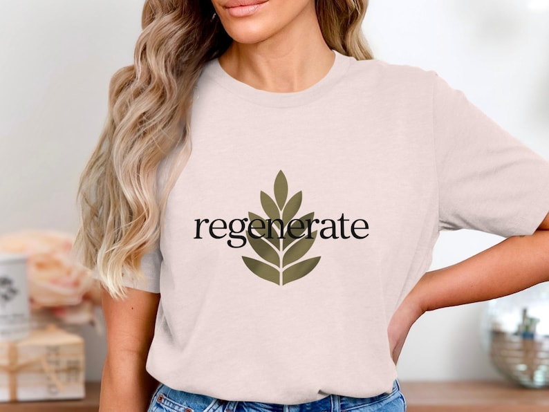 Puede incluir: Camiseta rosa claro con la palabra "regenerate" en escritura negra y un dise&ntilde;o de hojas verdes. La camiseta est&aacute; hecha de un material suave y es adecuada para ropa casual.