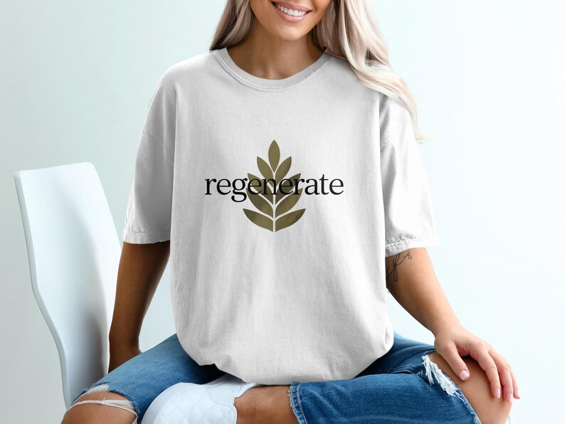 Puede incluir: Camiseta blanca con la palabra "regenerate" en negro y un dise&ntilde;o de hojas doradas. La camiseta tiene un corte holgado y oversize. La modelo lleva vaqueros azules.