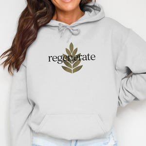 Puede incluir: Una sudadera con capucha gris claro con un bolsillo delantero y una capucha con cord&oacute;n. La palabra "regenerate" est&aacute; impresa en negro sobre un dise&ntilde;o de hojas verde oliva estilizado. La sudadera con capucha es de material suave.
