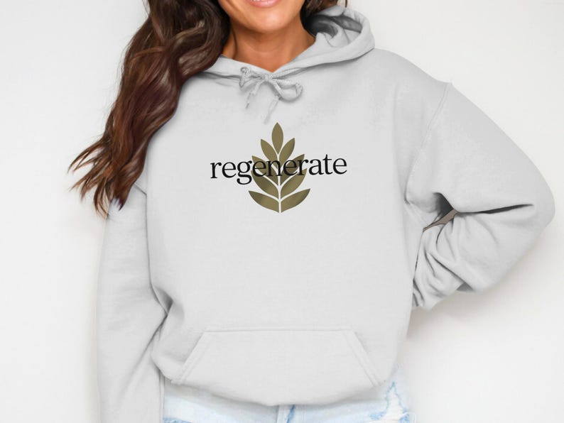 Puede incluir: Una sudadera con capucha gris claro con un bolsillo delantero y una capucha con cord&oacute;n. La palabra "regenerate" est&aacute; impresa en negro sobre un dise&ntilde;o de hojas verde oliva estilizado. La sudadera con capucha es de material suave.