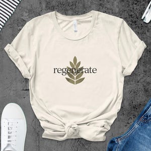 Puede incluir: Camiseta color crema con la palabra "regenerate" en fuente negra y un dise&ntilde;o de hojas. La camiseta se combina con zapatillas blancas y vaqueros azules. Tambi&eacute;n se ven un cuaderno y unos auriculares.