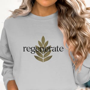 Puede incluir: Sudadera gris claro con la palabra "regenerate" en escritura negra y un dise&ntilde;o de hojas doradas. La sudadera es de cuello redondo y tiene mangas largas. El tema general es el bienestar y la naturaleza.