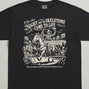 Op de afbeelding: Zwart t-shirt met een grafisch ontwerp met een skeletcowboy op een paard, met de tekst "It Was Also The Night That The Skeletons Came To Life." Het ontwerp bevat skeletten, gebouwen en extra tekst.
