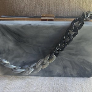 May include: A rectangular box clutch with a marbled gray and white acrylic finish. The bag features a gold-tone metal frame with a top clasp closure and a detachable chunky chain strap that transitions from light gray to solid black links.