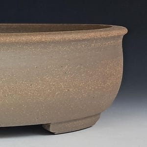 May include: A large, oval-shaped bonsai pot in a neutral, speckled tan color. The pot has a rounded rim and sits on three short, curved feet. The textured surface gives it a natural, earthy appearance, suitable for bonsai trees.