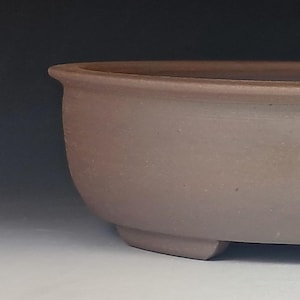May include: A shallow, oval-shaped bonsai pot crafted from unglazed, matte brown clay. The container features a smooth, curved exterior wall with a slightly flared rim at the top and is supported by small, integrated feet along the bottom base.