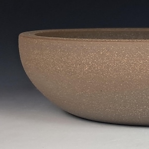 May include: A brown, oval-shaped ceramic planter with a textured surface. The planter has a low profile and a small drainage hole at the bottom. The neutral color and simple design make it suitable for various plants and decor styles.