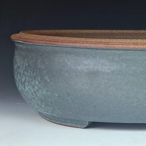 May include: An oval ceramic bonsai pot with a smooth, matte, sage green glaze on the exterior and a contrasting unglazed, textured, reddish-brown clay rim. The vessel features a low-profile, wide basin design with small feet at the base.