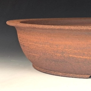 May include: A shallow, oval-shaped bonsai pot crafted from unglazed, reddish-brown terracotta clay. The vessel features a wide, flat rim, a smooth exterior surface with subtle horizontal striations, and a small arched cutout at the base.