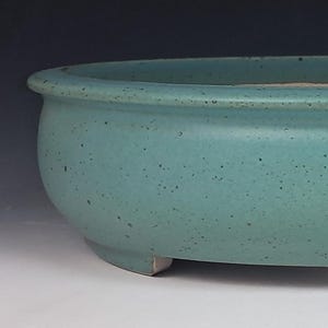 May include: A shallow, oval-shaped ceramic bonsai pot with a matte, speckled teal-green glaze. The container features a wide, rounded rim and rests on small, integrated feet, showcasing a smooth, uniform texture across its exterior surface.