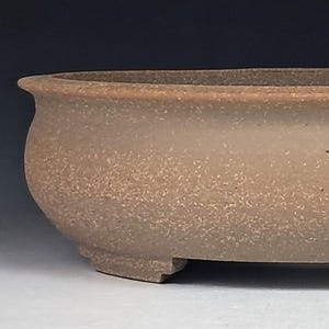 May include: A low, oval-shaped ceramic bonsai pot with a textured, matte tan finish. The vessel features a wide, slightly flared rim and rests on four small, integrated feet. The exterior surface displays a speckled, earthy appearance.