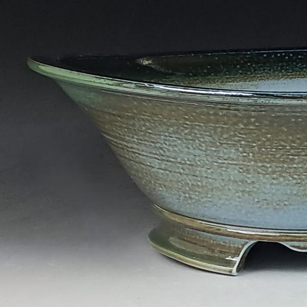 May include: A handcrafted ceramic bowl with a wide, shallow design. The bowl features a textured surface with a gradient of colors, including shades of blue, green, and brown. It rests on a low, three-footed base, showcasing a unique pottery piece.