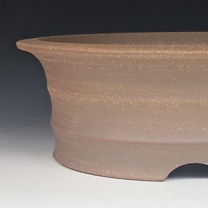 May include: A round, unglazed brown ceramic bonsai pot with a wide, flared rim and a textured, matte finish. The vessel features a subtle horizontal ridged pattern along its exterior walls and a single arched drainage hole at the base.