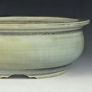 May include: A round, light green ceramic bonsai pot with a wide, shallow design. The pot has a slightly flared rim and a rounded body, with a dark gray band around the top edge. The base has three drainage holes.