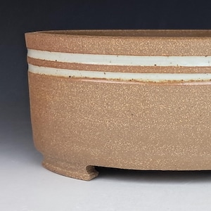 May include: A handcrafted, oval-shaped ceramic planter. The planter is a speckled tan color with two horizontal white bands near the top. It has a textured surface and sits on four small feet. Ideal for succulents or small plants.