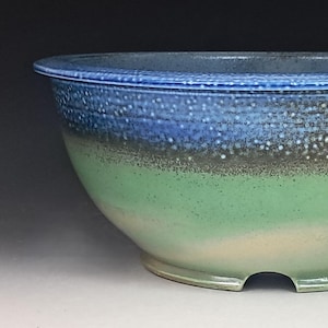 May include: A ceramic bowl featuring a gradient glaze that transitions from a deep blue rim with speckled white details to a muted green base. The bowl has a wide, rounded opening and a footed bottom with a small arched cutout notch.