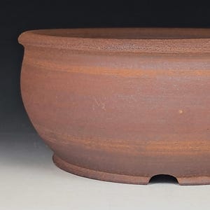 May include: A round, unglazed terracotta bonsai pot with a matte, earthy brown finish. The vessel features a wide, slightly curved body, a thick rolled rim at the top, and a small drainage notch cut into the base of the footed bottom edge.