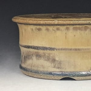 May include: A round ceramic bonsai pot with a wide, flared rim and a cylindrical body. The exterior features a matte tan finish with dark brown, weathered-looking horizontal streaks and a dark, glossy blue-black glazed band at the base.