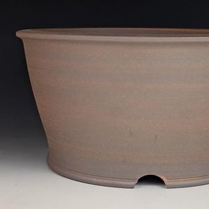 May include: A large, brown ceramic bonsai pot with a wide, oval shape. The pot has a slightly textured surface and a drainage hole at the base. The rim is rounded, and the sides gently slope inward.