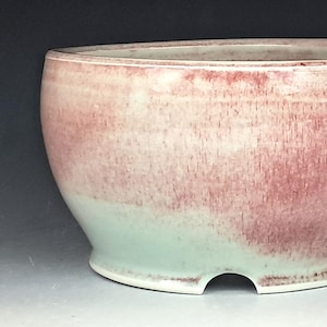 May include: A handmade ceramic bowl with a unique design. The bowl features a gradient of colors, transitioning from a soft mint green at the base to a dusty rose at the top. The bowl has a rounded shape with a flat base and a small cutout.