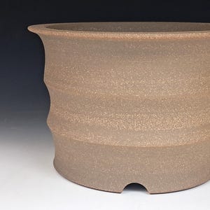 May include: A cylindrical ceramic planter with a speckled light brown finish. The exterior features three horizontal ridges that create a wavy profile. The top rim is flared outward, and the base includes a small arched drainage cutout.