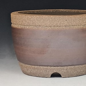 May include: A round ceramic planter featuring a smooth, dark brown central band contrasted by a textured, light brown rim and base. The pot has a wide opening and a small, arched drainage notch cut into the bottom edge of the base.