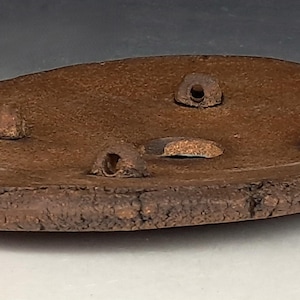 May include: A handmade, oval-shaped, brown ceramic dish with a rough, textured edge. The dish features four small, cone-shaped protrusions and a central, rectangular opening. The overall design suggests a rustic, artisanal aesthetic.