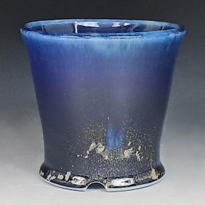 May include: A ceramic cup featuring a deep blue glaze that transitions into a darker, near-black base. The surface displays a mottled, textured finish near the bottom with metallic-looking flecks and a small, arched cutout at the base rim.