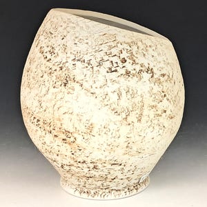 May include: A spherical ceramic vase with a wide, slanted opening at the top. The exterior surface features a textured, off-white finish with mottled brown speckles and irregular, rough patches distributed across the rounded body and base.