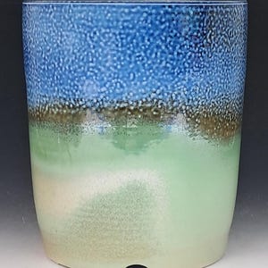 May include: A cylindrical ceramic planter featuring a gradient glaze that transitions from a textured, deep blue at the top to a smooth, light mint green and cream at the base, with a small arched cutout at the bottom edge of the vessel.