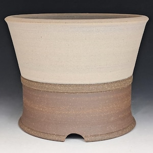May include: A ceramic planter with a wide, flared top section in a matte off-white finish and a cylindrical base in a textured, earthy brown tone. The base features a small, semi-circular cutout at the bottom edge, providing a distinct two-tone look.