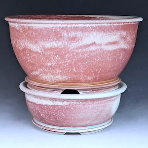 May include: Two stacked, reddish-pink ceramic planters with a textured, weathered appearance. The larger planter sits atop a smaller one, both featuring a rounded shape and a white rim. Ideal for indoor or outdoor plants.